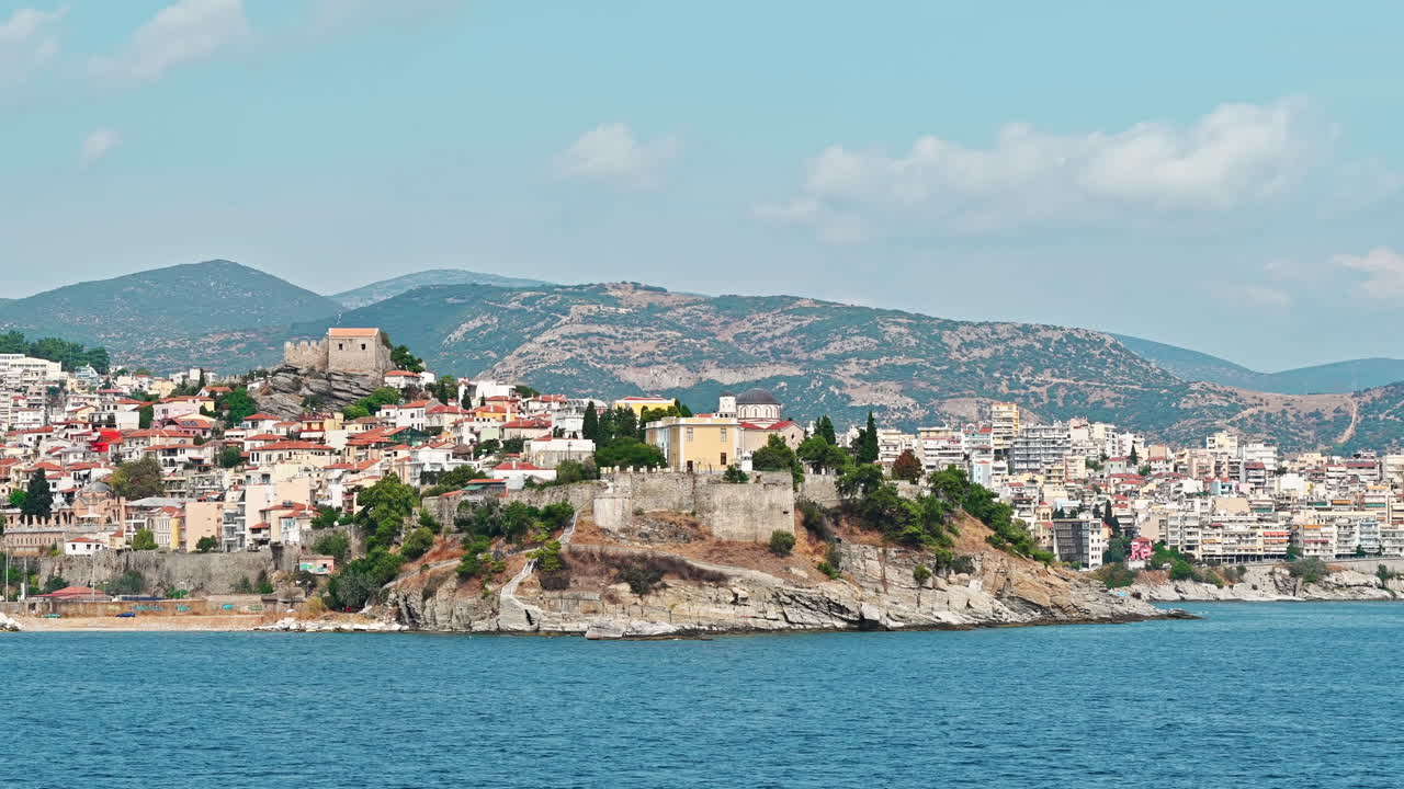 View of Kavala from the water. Rows of residential buildings, ancient fortress and church on the hill, Aegean sea rocky coast. Greece