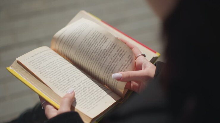 Close-up of a woman s hands turning a page of her book outdoors, nails painted with white polish, yellow-edged book open, showing text in the pages