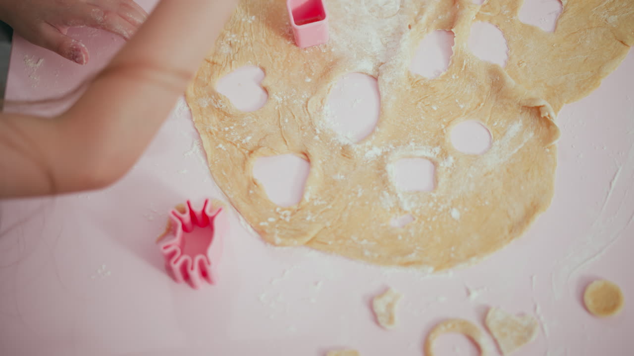 Top down view of fair skinned child cutting rolled dough using pink shape cutter on pink surface, creating multiple heart and circle shapes during fun baking activity with flour scattered around