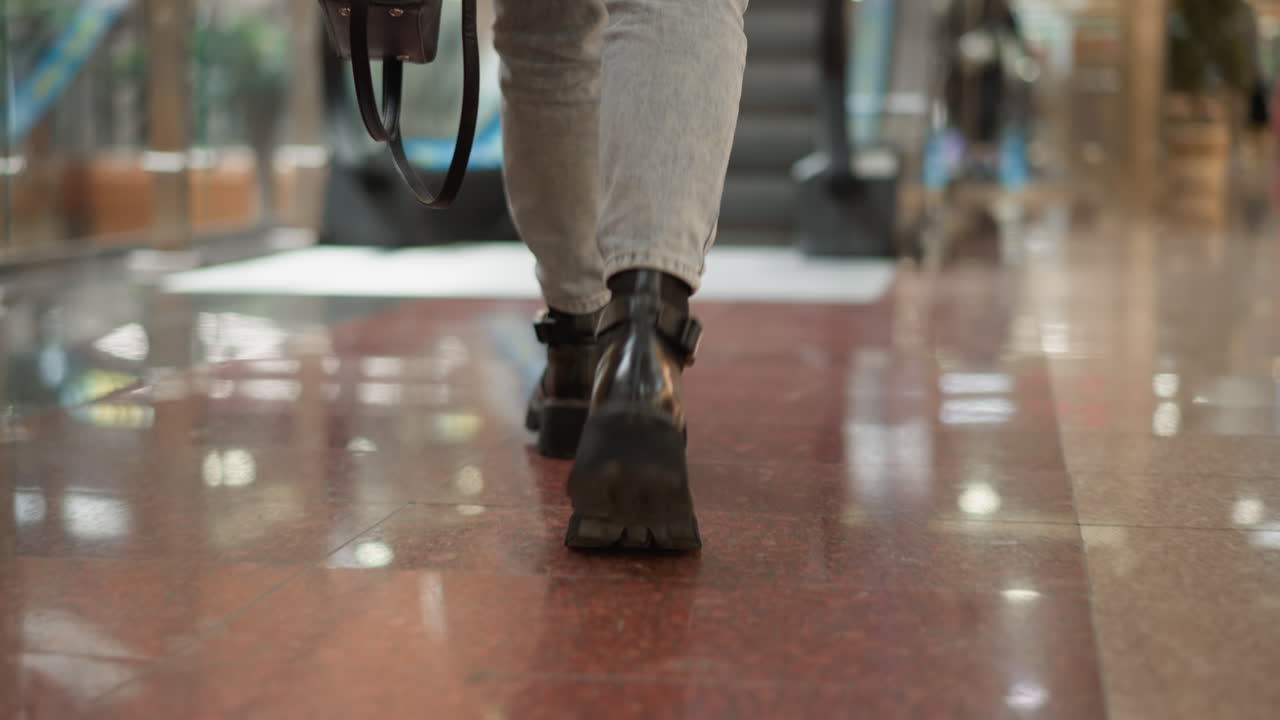 high angle intimate leg view of modern woman wearing jeans and black boots carrying bag walking on glossy tiled floor toward moving walkway inside bright retail mall interior next to glass railing