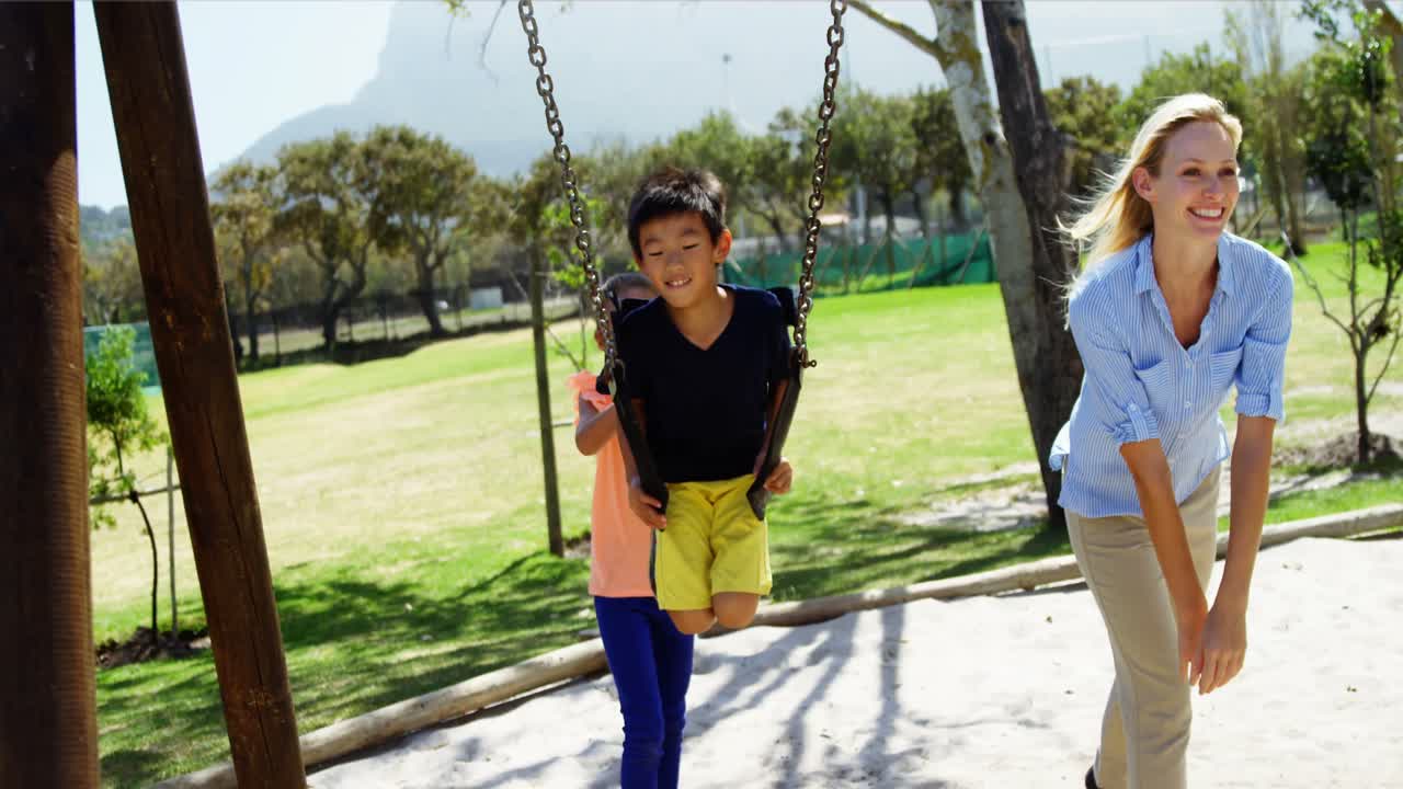 entrenador feliz empujando a los escolares en el columpio en el patio de recreo