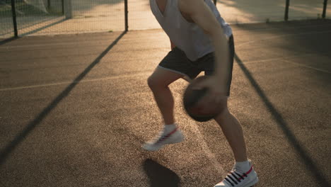 Ein-Männlicher-Basketballspieler,-Der-Den-Ball-Bei-Sonnenuntergang-Auf-Einem-Außenplatz-Zwischen-Seinen-Beinen-Hüpft-Und-Dribbelt-1