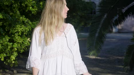 A-happy-blonde-woman-in-a-white-dress-walking-and-enjoying-music-in-the-summer-park,-smiling