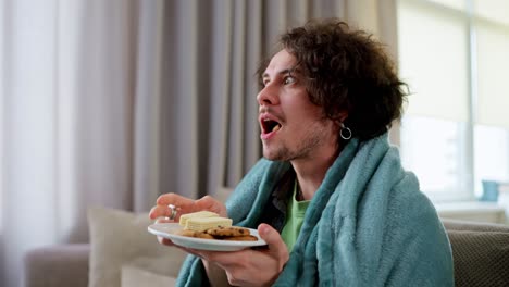 Happy-interested-brunette-guy-with-curly-hair-eats-sweet-snacks-and-watches-incredible-movies-while-sitting-on-the-couch-at-home-wrapped-in-a-blanket