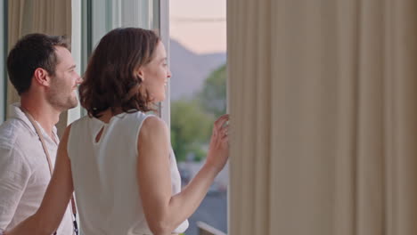 happy-travel-couple-arriving-in-hotel-room-on-vacation-looking-out-window-young-man-photographing-view-enjoying-romantic-holiday-with-wife