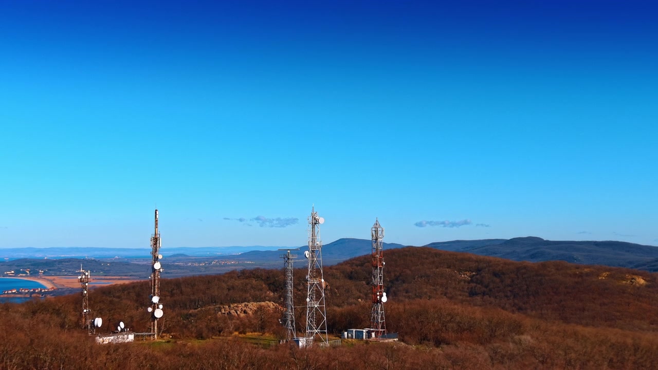 Veduta aerea di antenne di comunicazione su una cresta montuosa. Molteplici torri d'acciaio con parabole satellitari sopra un bosco spoglio, mare visibile all'orizzonte.
