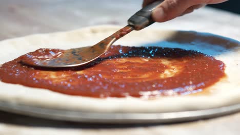 a close-up of a pizza dough on a steel tray on wooden counter while an expereinced chef put juicy red sauce on it with a adicid