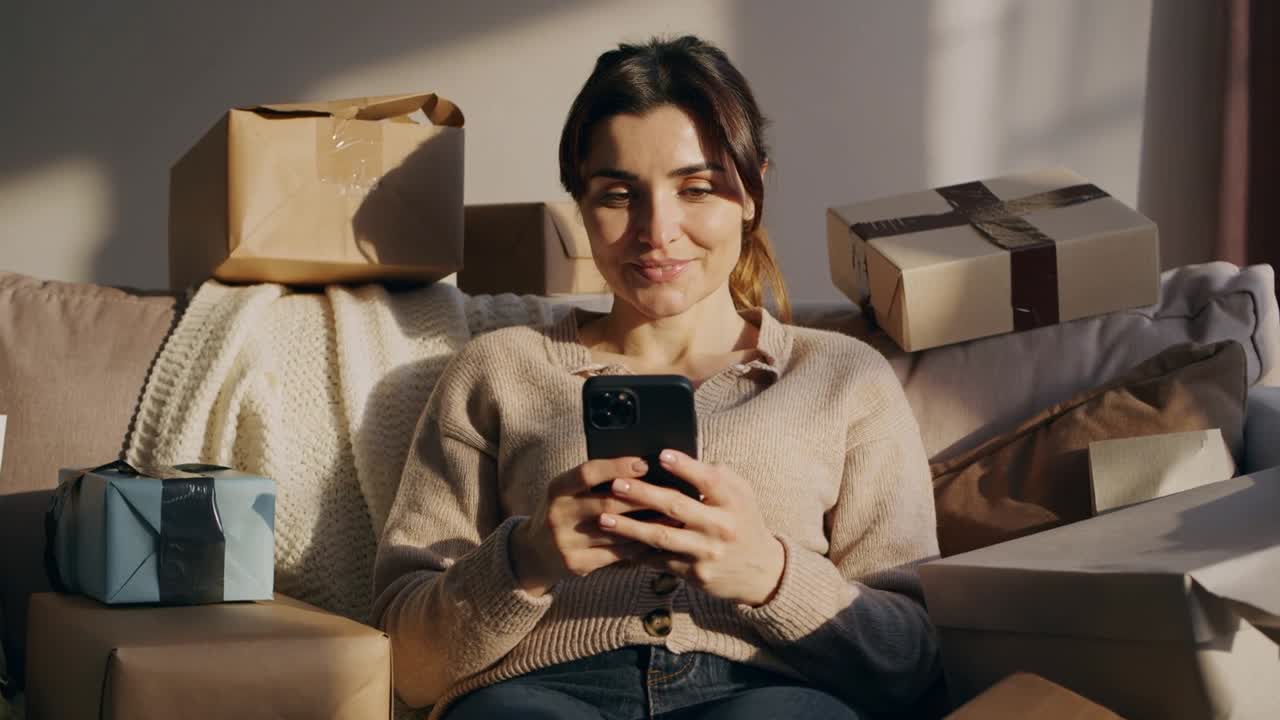 Warm sunlight motivating woman raising smartphone, scrolling tapping on sofa at home with packages