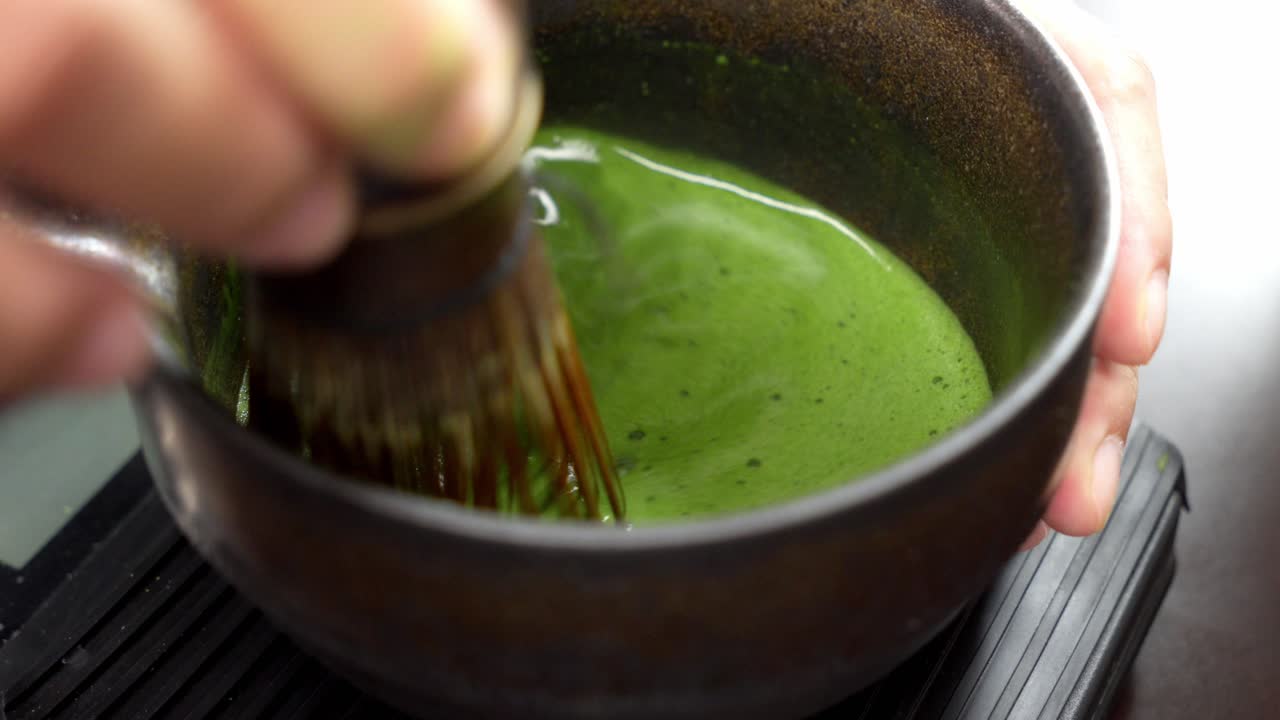 Close-up de um batedor de bambu sendo usado para preparar chá matcha verde cerimonial na Indonésia. O movimento cria uma rica espuma verde em uma tigela de cerâmica tradicional