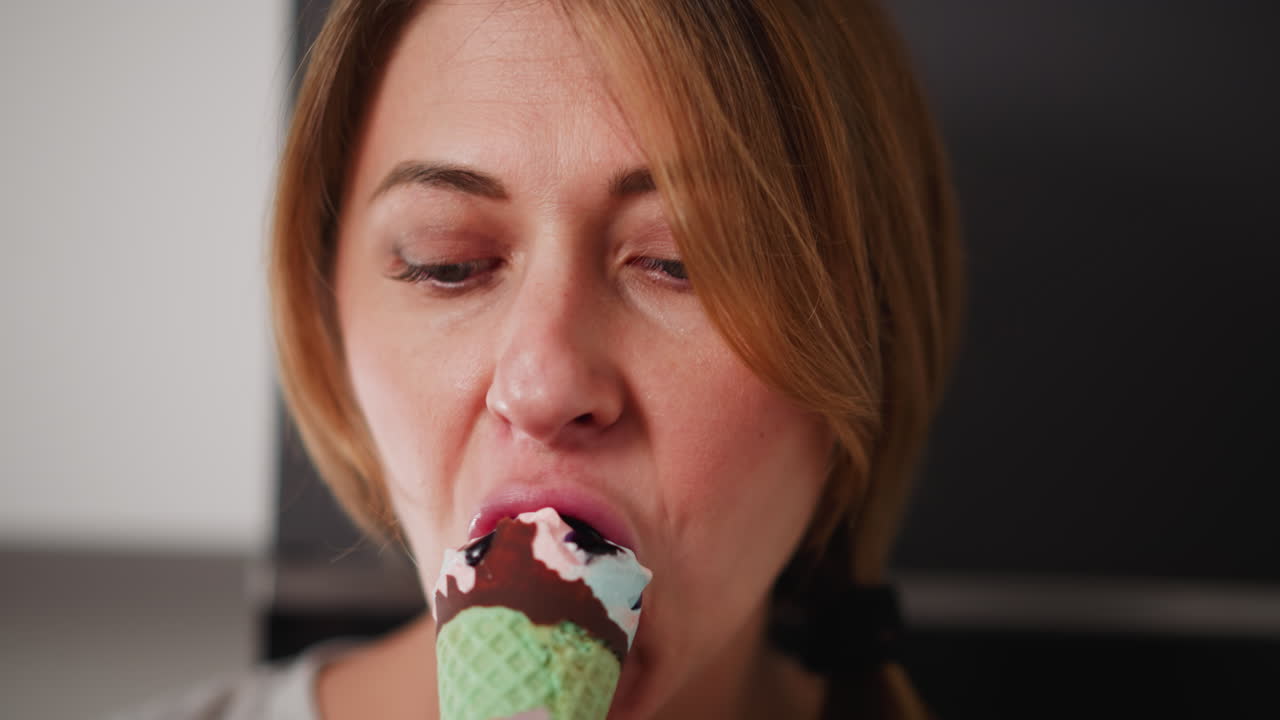 Close up of lady savoring sweet dessert holding colorful green cone, enjoying taste with visible creamy texture on lips, blurred kitchen background