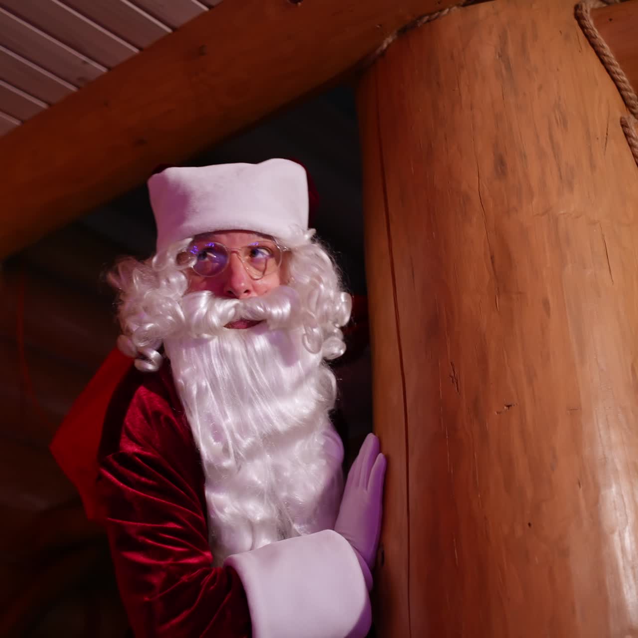 Santa Claus hiding in a wooden house. Authentic Santa with white beard and red costume with a sack with presents looking out of wooden pillar