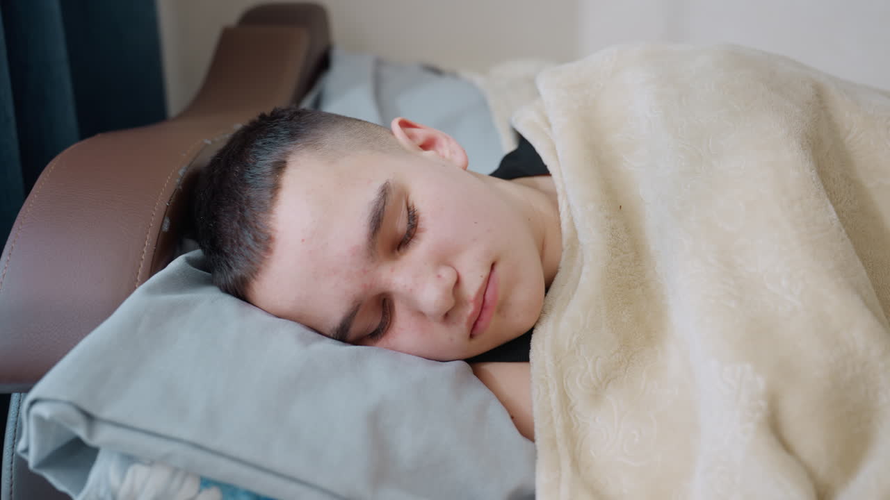 Little boy sleeping peacefully under blanket on bed with gentle sunlight streaming through window and blue curtain creating warm serene cozy atmosphere of childhood rest