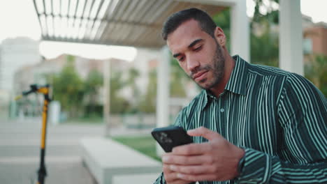 Junger-Mann-Schreibt-SMS-Auf-Dem-Smartphone-Und-Sitzt-Draußen-Auf-Einer-Bank.