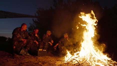 a large bonfire flame, behind it military men in camouflage uniforms are sitting, talking and warming themselves at night by the hearth