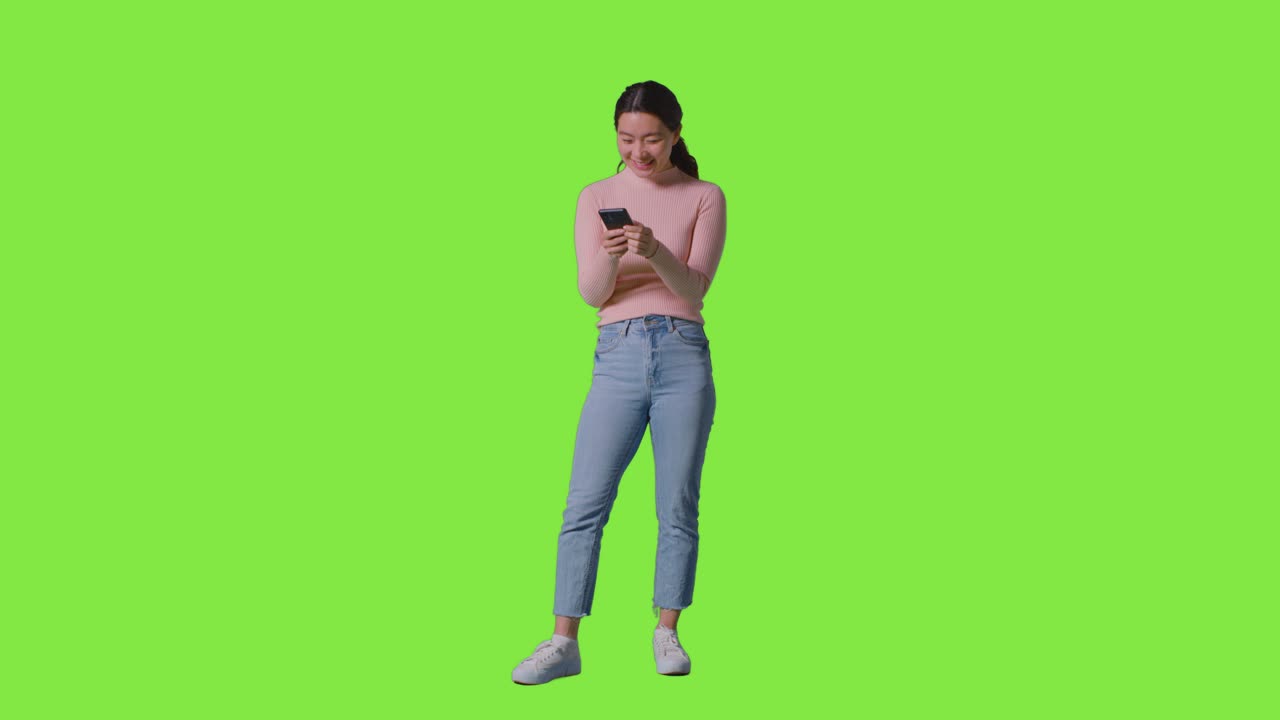 Full Length Studio Shot Of Woman Smiling At Message Or Content On Mobile Phone Against Green Screen