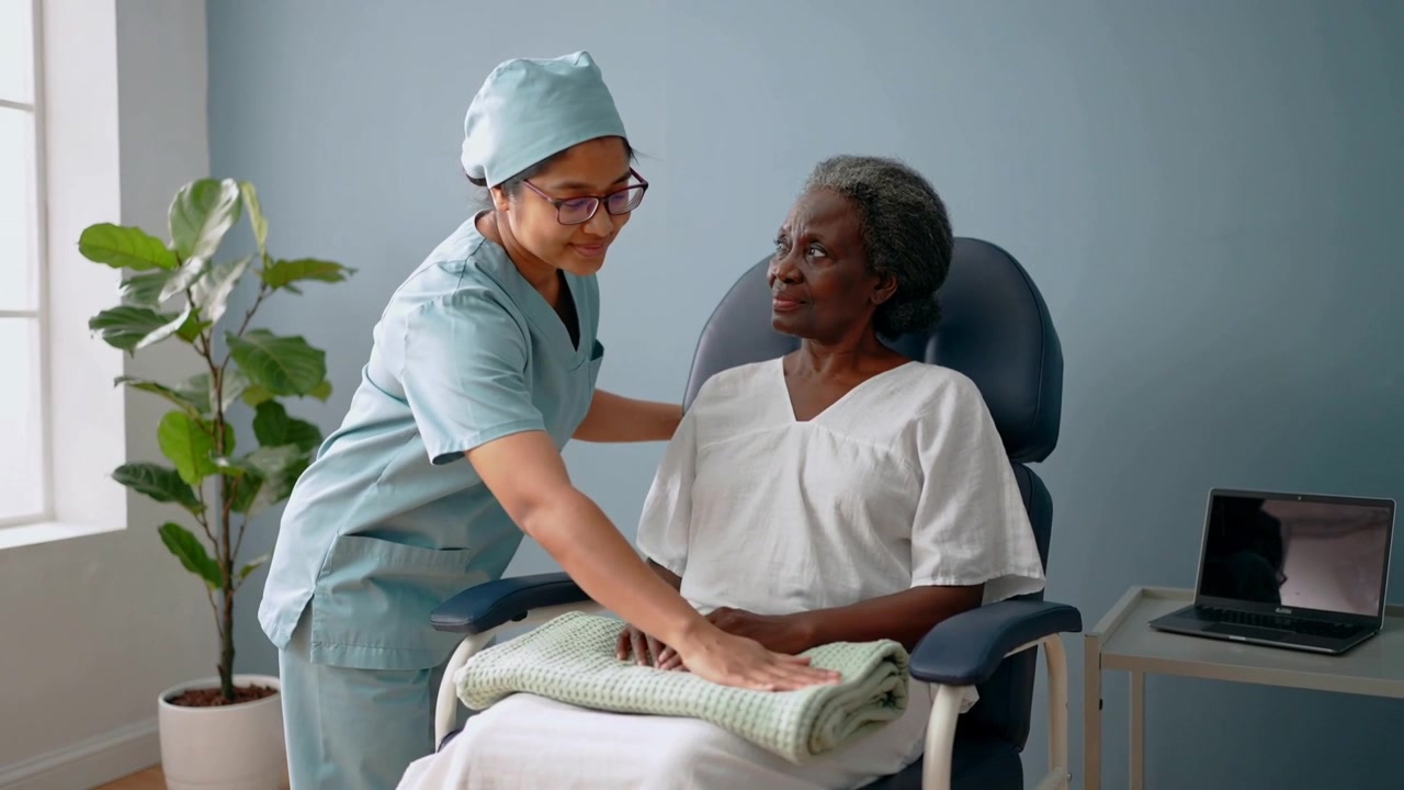 Noticing patient in gown, caregiver in scrubs placing blanket smoothing lap in clinic offering aid