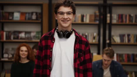 close up portrait cheerful young man laughing enjoying successful lifestyle achievement. european male in plaid shirt and headphones in library. bookshelf and classmates on background college education