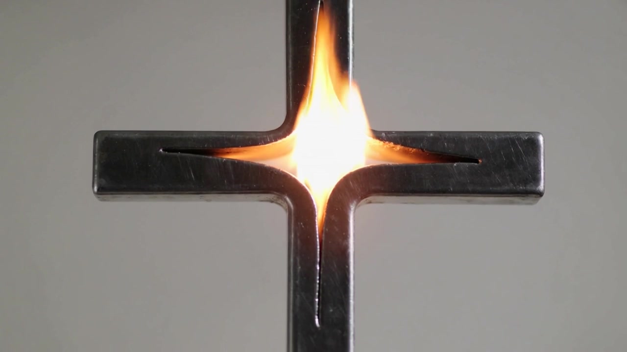Croce metallica scintillante che emette una fiamma che sale da una cavità centrale su uno sfondo grigio, mostrando fuliggine.