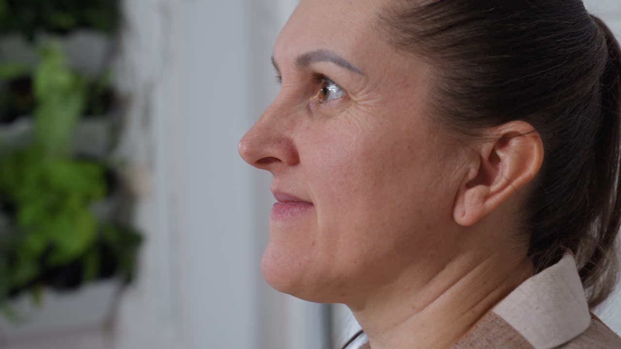 Cheerful woman smiles softly while gazing upward in bright indoor garden setting, blurred green foliage behind adds serene natural atmosphere to moment of mindful plant care and relaxed enjoyment