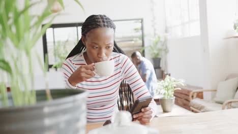 Fröhliche-Afroamerikanische-Frau,-Die-Ihr-Smartphone-Benutzt-Und-Kaffee-Am-Tisch-Im-Café-Trinkt,-Zeitlupe