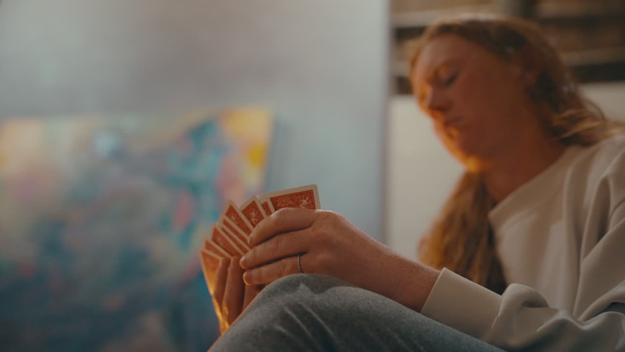 Woman sitting fireside holding red hand of cards reaches to draw another, wide handheld 4k