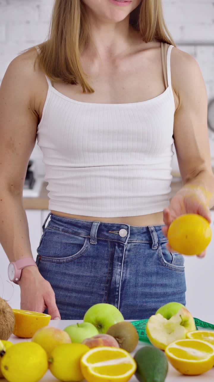 Young woman with muscular body in the kitchen. Sporty female juggling with fresh fruit on the background of healthy food. Healthy diet concept. Vertical video