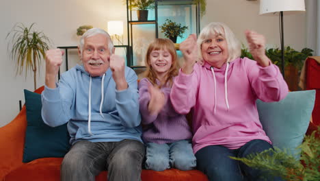 happy caucasian grandparents child girl granddaughter watching tv celebrating success winning goal