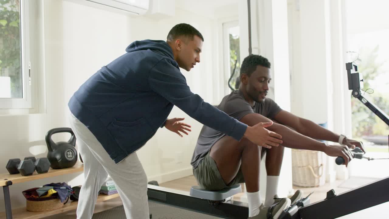 Male trainer entering home gym guiding African American client on rowing machine for proper form