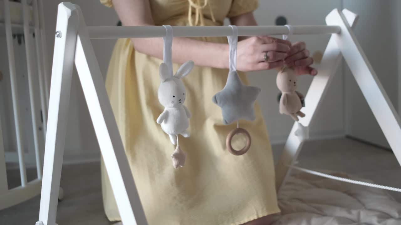Expecting mom hanging stuffed animal toys on white wooden baby play gym in nursery