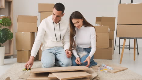 Front-View-Of-A-Young-Couple-In-A-New-House-Sitting-On-The-Carpet-Assembling-A-Furniture-1