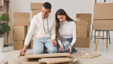 front view of a young couple in a new house sitting on the carpet assembling a furniture 1