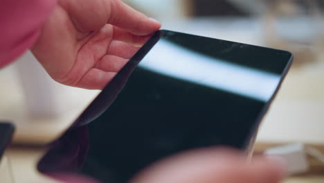 lady holding a tablet from its stand in an electronics store, connected by a security cord, she gently inspects the device while light reflects off the screen, with a blur background