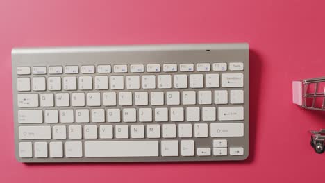 overhead view of computer keyboard, shopping trolley on pink background