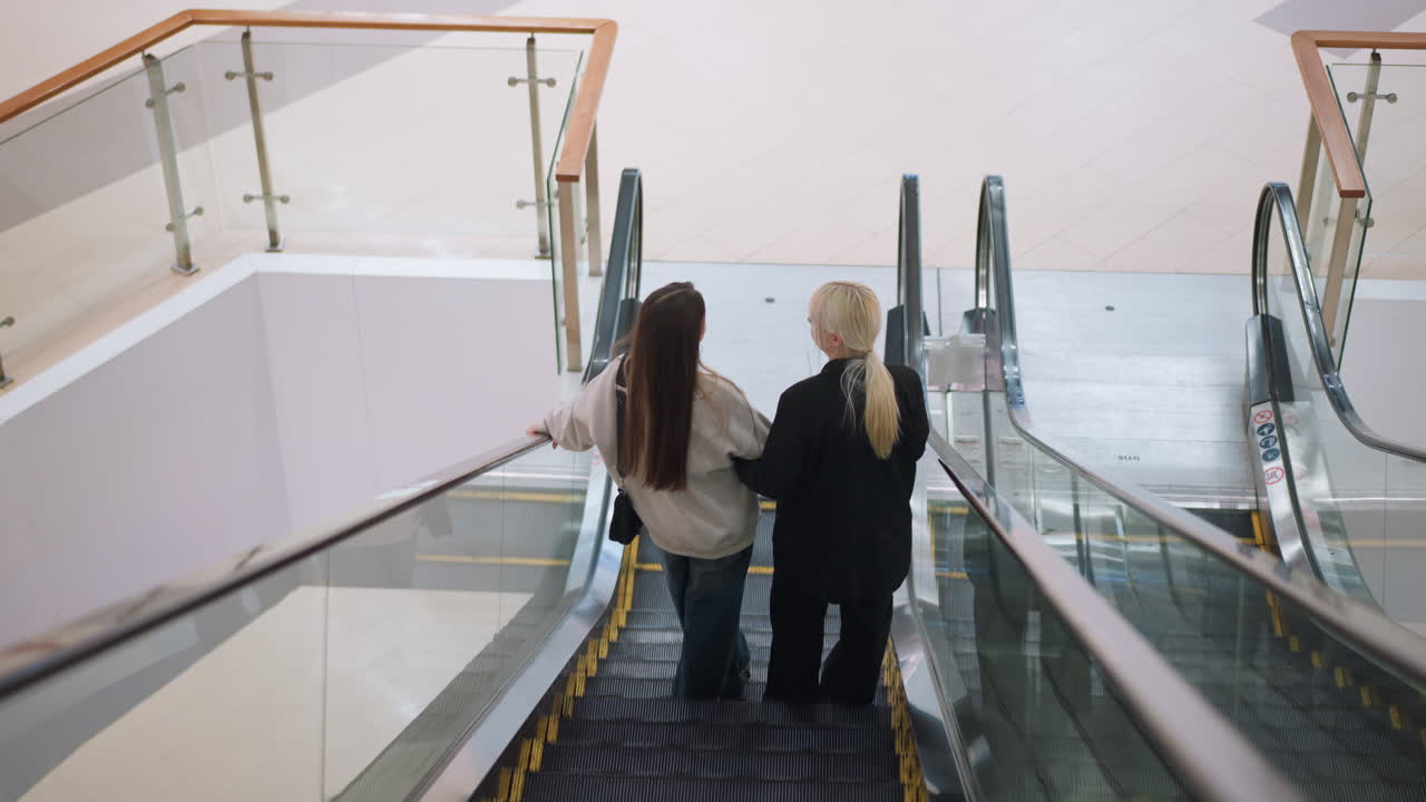 Back view of two women descending on escalator in modern shopping mall, holding drink and chatting while enjoying leisure time together, highlighting friendship, lifestyle