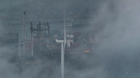 aerial shot of a wind turbine and dense industrial center through the clouds near the port of vlissingen with a ship in the harbor before being obscured