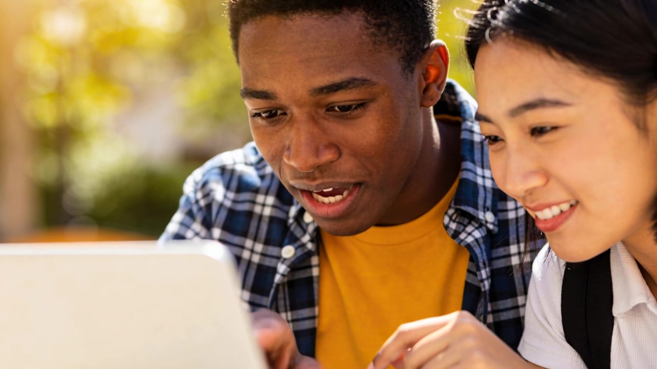 Leaning pair using laptop at park bench, man pointing at screen, laughing then focusing, backpack
