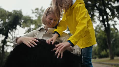 cerca de una feliz mujer rubia en una chaqueta blanca acariciando a su gran perro negro con su hija mientras caminaba en el parque después de la lluvia