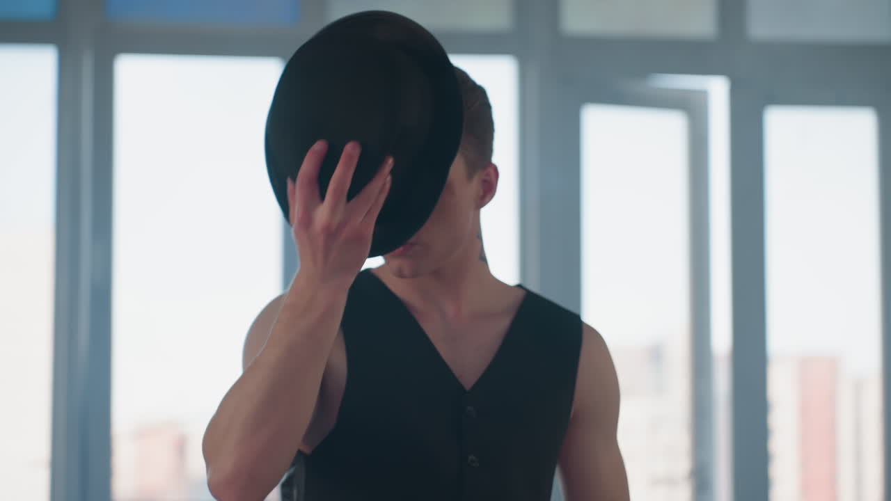 handsome man in vest and black pants dances in sunlit studio removes hat touches hair then replaces hat with smooth movement against tall glass windows with smoke swirl framing urban skyline