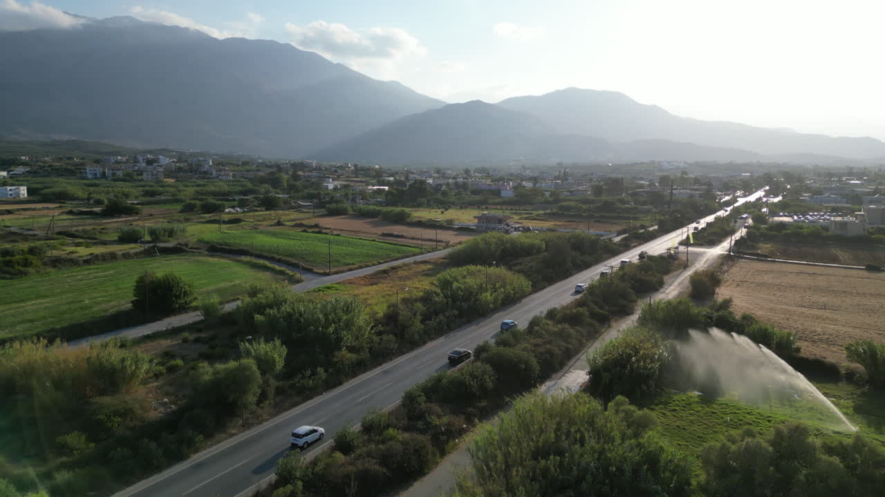 드론은 눈에 띄는 자동차가 있는 바쁜 도로 위를 날아다니며, 전면에  관개와 멀리서 높은 산을 보여 주고, 밝은  구름을 통해 빛이 비추고 있습니다.