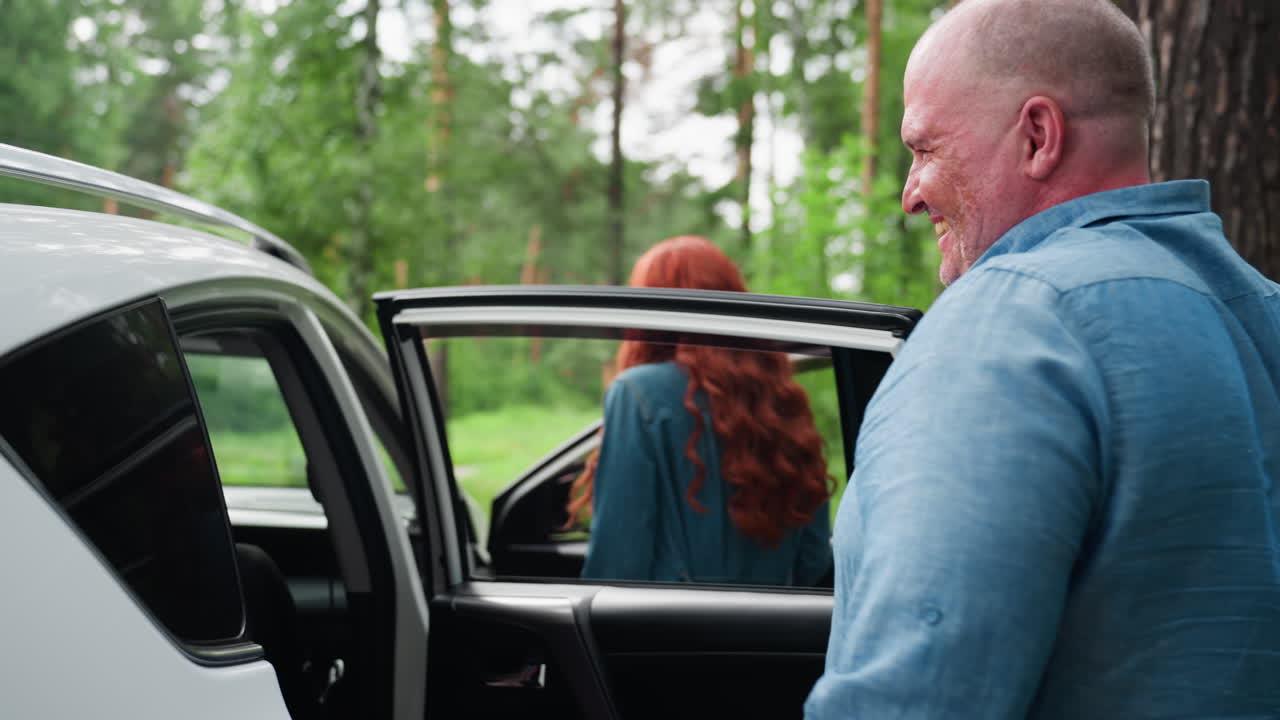 Back view of head of family smiling while closing back door of white car parked in lush green forest, enjoying calm atmosphere surrounded by nature, trees, and sunlight reflecting on vehicle surface