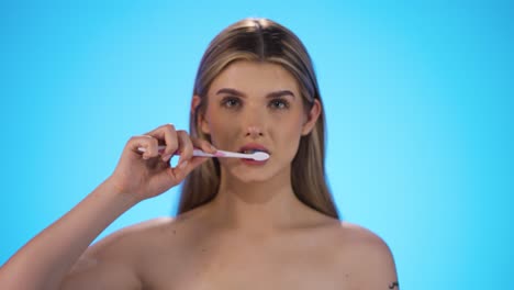 Statische-Zeitlupenaufnahme-Einer-Jungen-Hübschen-Blonden-Frau,-Die-Eine-Zahnbürste-In-Die-Kamera-Hält-Und-Sich-Dann-Die-Zähne-Putzt,-Um-Eine-Gute-Tägliche-Zahnpflege-Zu-Gewährleisten-Und-Karies-Vor-Blauem-Hintergrund-Vorzubeugen