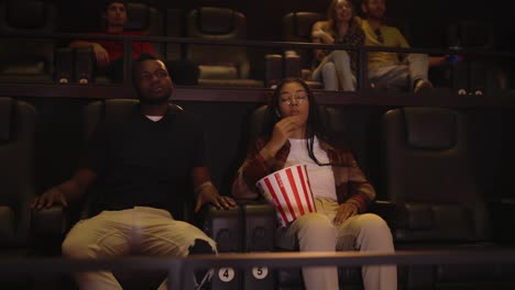 african american young couple sitting in movie theatre watching film and taking popcorn