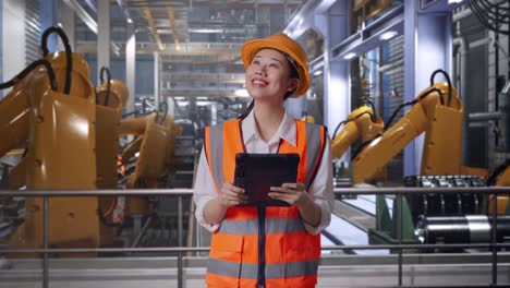 asian female engineer with safety helmet looking at the tablet in her hand and looking around while standing with industrial robotic arms working