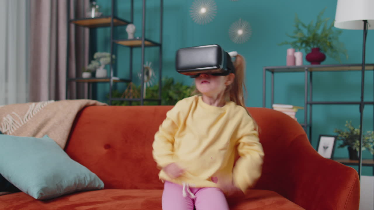 Toddler girl sitting on home sofa using virtual reality headset helmet app to play video 3D game
