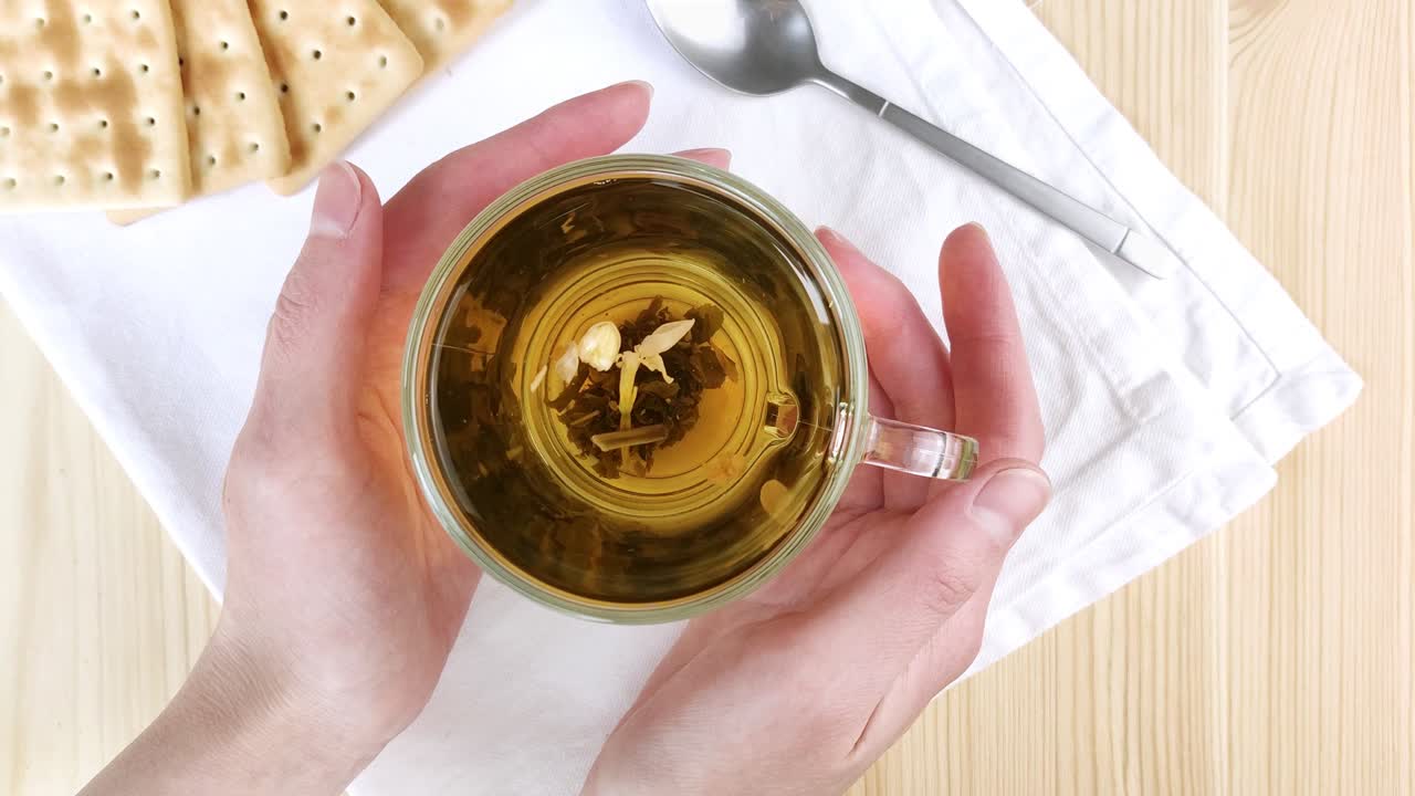Green tea with jasmine is brewed, leaves move in cup in circle. Female hands hold hot drink in glass