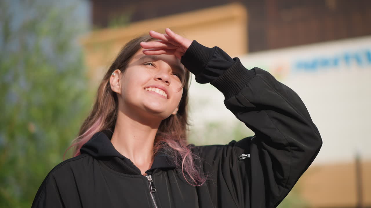 Adolescente mira hacia arriba después de la lluvia. Joven sonriente protegiéndose los ojos al aire libre. Un adolescente con cabello rosa húmedo bajo un cielo brillante. Joven con cabello rosa mira felizmente al cielo después de la lluvia.