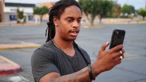 hombre afroamericano con rastas y tatuajes saca el teléfono para hacer una videollamada en el estacionamiento