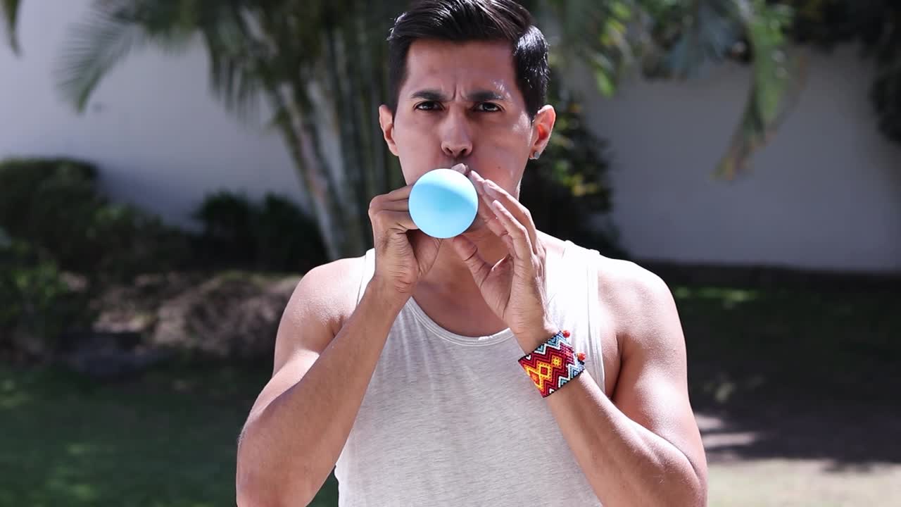 심각한 젊은 수염 없는 남자가 열대지방에서 라텍스 풍선을 부풀리고 있습니다.