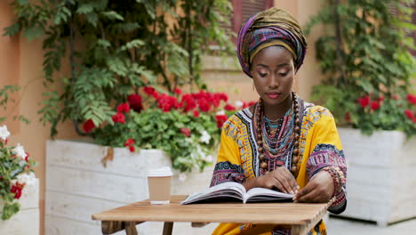 Mujer-Joven-Atractiva-Y-Elegante-Con-Ropa-Tradicional-Sentada-En-La-Mesa-En-El-Hermoso-Patio-Exterior-Con-Flores,-Leyendo-Un-Libro-Y-Tomando-Un-Café-O-Té