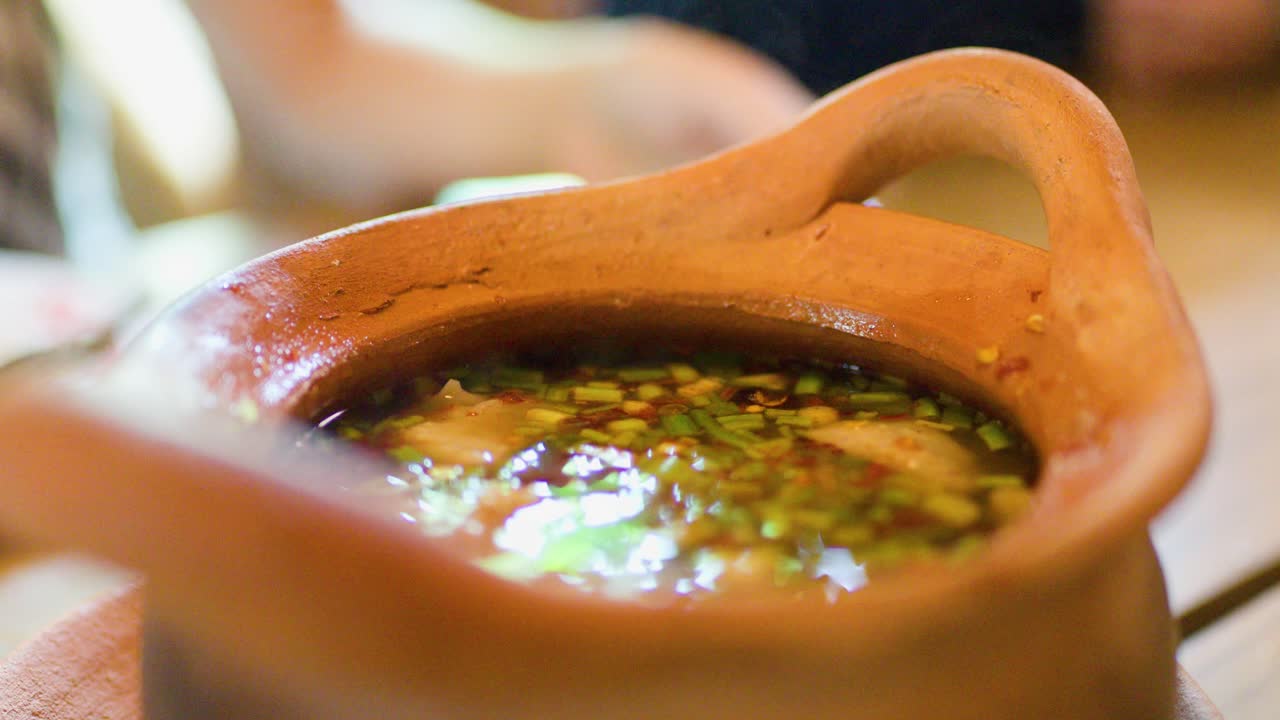 Hand lifts clay pot lid, unveiling steaming Thai herbal soup in warm, natural lighting